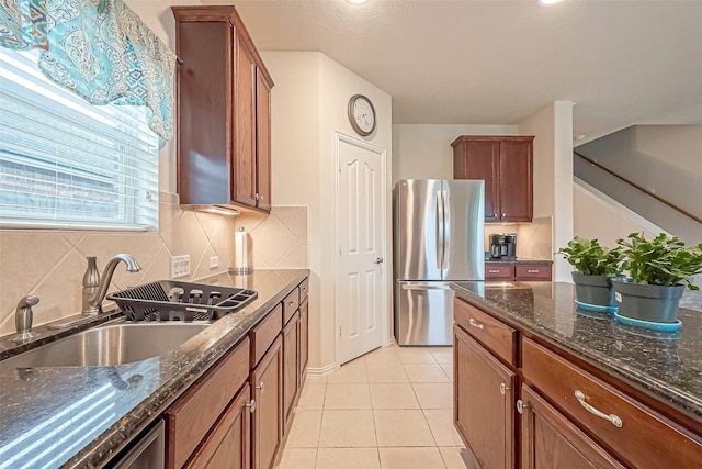 kitchen with brown cabinets, freestanding refrigerator, light tile patterned flooring, a sink, and dark stone counters