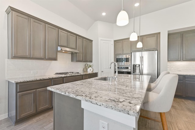 kitchen with a center island with sink, appliances with stainless steel finishes, light stone counters, under cabinet range hood, and a sink