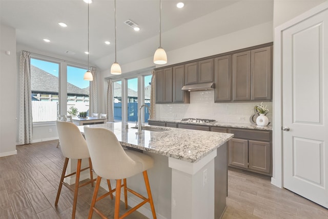 kitchen with light wood-style flooring, a kitchen island with sink, under cabinet range hood, a sink, and backsplash