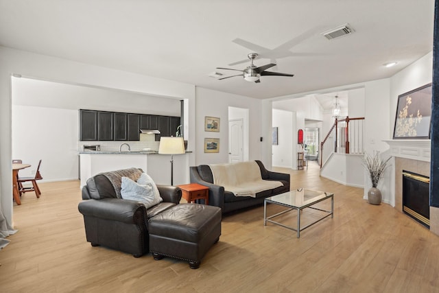 living area with visible vents, stairway, light wood-style floors, a fireplace, and ceiling fan with notable chandelier