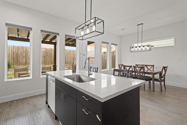 kitchen featuring a center island with sink, dishwashing machine, dark cabinets, light countertops, and a sink