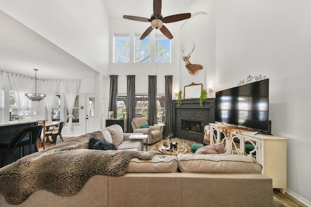 living area featuring a towering ceiling, a stone fireplace, wood finished floors, baseboards, and ceiling fan with notable chandelier