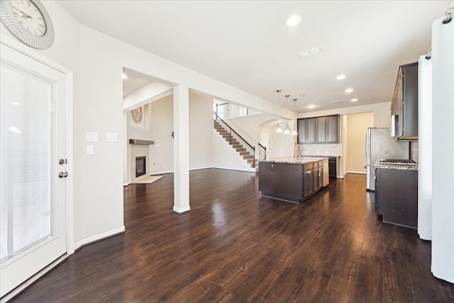 kitchen featuring dark brown cabinetry, dark wood finished floors, a tiled fireplace, an island with sink, and backsplash