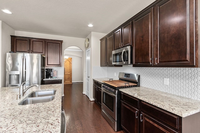 kitchen featuring arched walkways, stainless steel appliances, a sink, dark brown cabinets, and light stone countertops