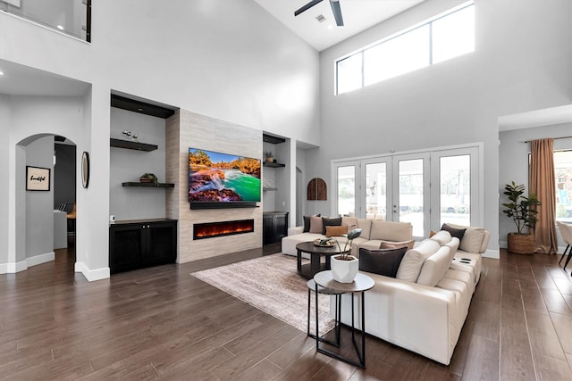 living room featuring arched walkways, built in shelves, dark wood-style flooring, baseboards, and a lit fireplace