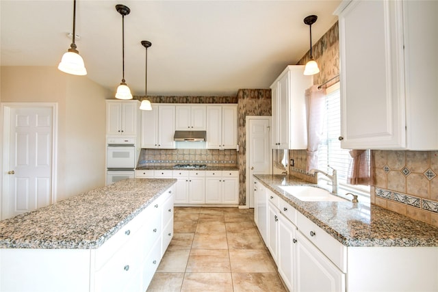 kitchen with white double oven, under cabinet range hood, stainless steel gas cooktop, a kitchen island, and a sink