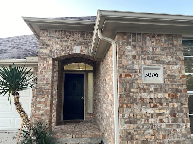 doorway to property with an attached garage, brick siding, and roof with shingles