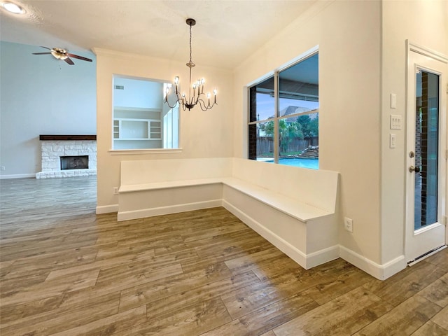 unfurnished dining area featuring a stone fireplace, ceiling fan with notable chandelier, wood finished floors, baseboards, and crown molding