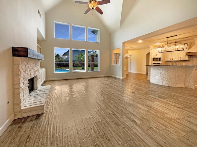 unfurnished living room with light wood-style floors, a sink, a stone fireplace, baseboards, and ceiling fan with notable chandelier