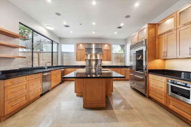 kitchen featuring appliances with stainless steel finishes, wall chimney range hood, a kitchen island with sink, and finished concrete floors