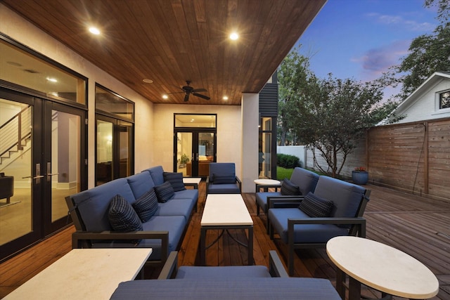 patio terrace at dusk with a deck, fence, an outdoor living space, a ceiling fan, and french doors