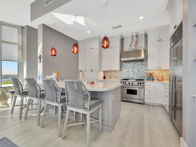 kitchen featuring high end appliances, tasteful backsplash, light countertops, light wood-style flooring, and wall chimney range hood