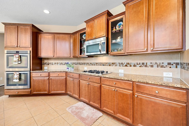 kitchen with light tile patterned floors, decorative backsplash, appliances with stainless steel finishes, brown cabinets, and light stone counters