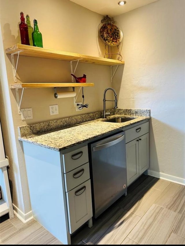 kitchen with light stone counters, gray cabinetry, stainless steel dishwasher, open shelves, and a sink