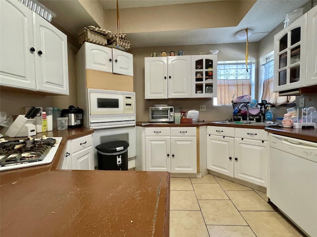 kitchen with light tile patterned floors, white appliances, a sink, dark countertops, and glass insert cabinets