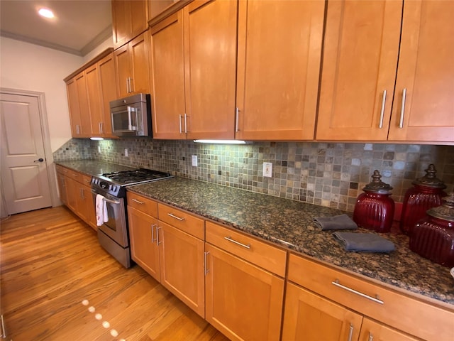 kitchen with appliances with stainless steel finishes, dark stone counters, crown molding, and light wood-style flooring