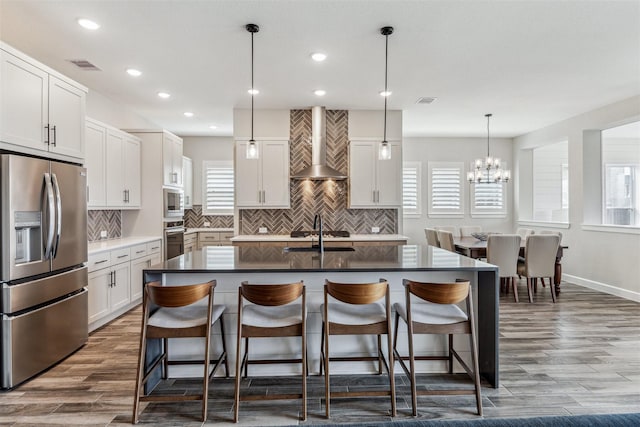 kitchen with stainless steel appliances, a kitchen bar, a kitchen island with sink, a sink, and wall chimney exhaust hood