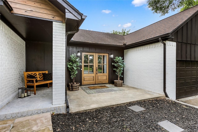 view of exterior entry featuring a shingled roof, an attached garage, french doors, board and batten siding, and brick siding