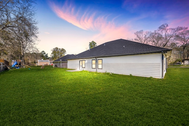 back of house at dusk featuring a shingled roof, a playground, and a yard