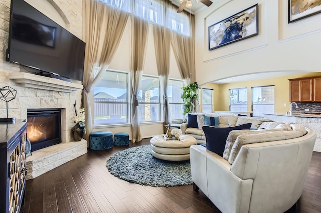 living room featuring dark wood-style floors, a stone fireplace, a high ceiling, and a ceiling fan