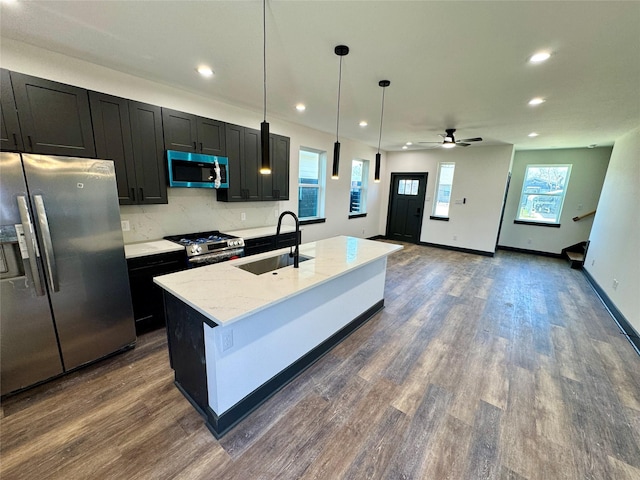 kitchen with dark wood finished floors, appliances with stainless steel finishes, dark cabinets, a kitchen island with sink, and a sink