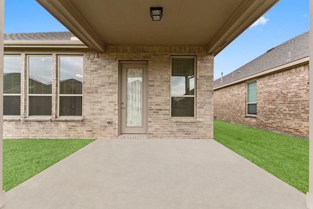 property entrance featuring a patio, brick siding, a lawn, and roof with shingles