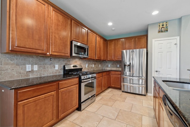 kitchen featuring decorative backsplash, brown cabinetry, appliances with stainless steel finishes, dark stone countertops, and a sink