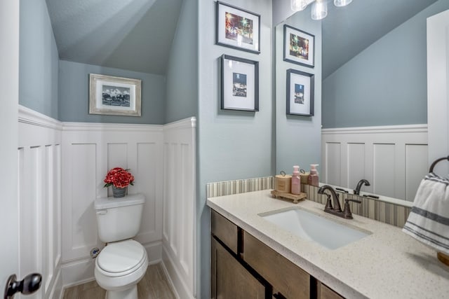 half bath featuring toilet, a wainscoted wall, wood finished floors, vanity, and vaulted ceiling