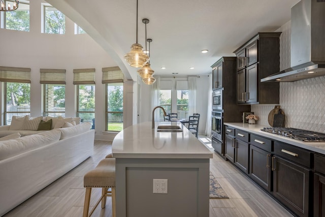 kitchen with open floor plan, stainless steel appliances, light countertops, wall chimney range hood, and a sink