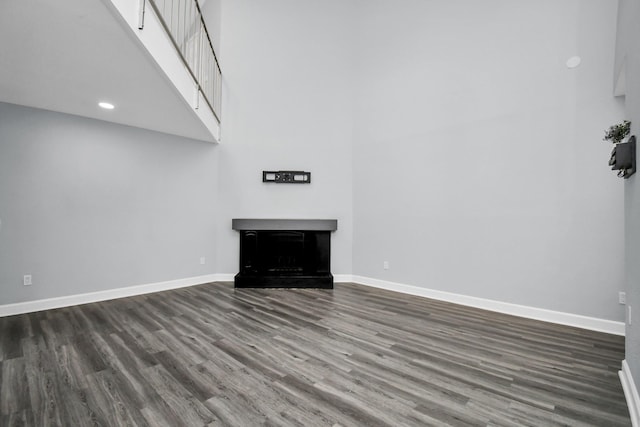 unfurnished living room with dark wood-style flooring, recessed lighting, a fireplace with raised hearth, a high ceiling, and baseboards