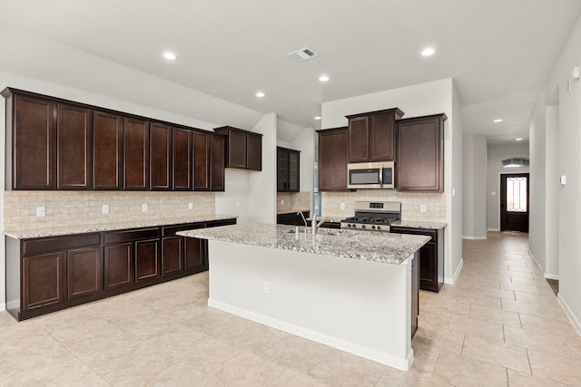 kitchen with light stone counters, a kitchen island with sink, stainless steel appliances, visible vents, and dark brown cabinets