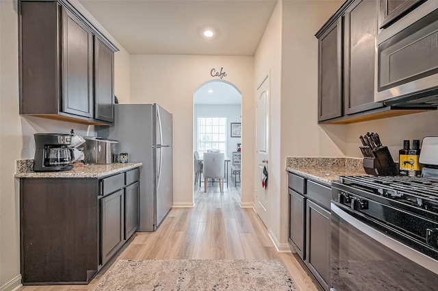 kitchen featuring arched walkways, light wood finished floors, appliances with stainless steel finishes, dark brown cabinetry, and light stone countertops