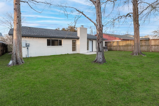 back of house featuring roof with shingles, brick siding, a chimney, a lawn, and a fenced backyard