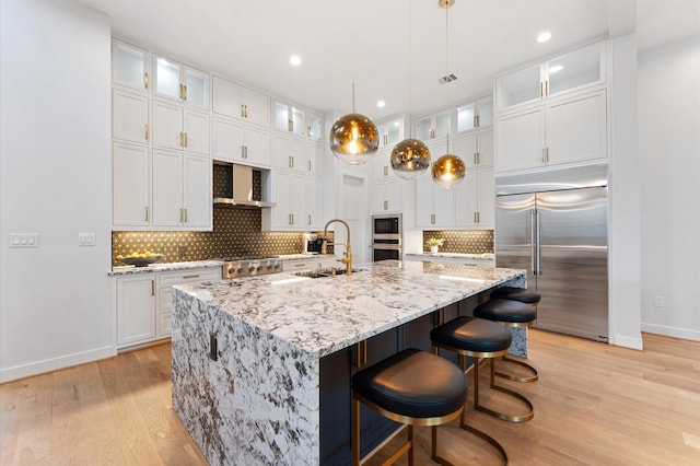 kitchen with white cabinetry, wall chimney range hood, built in appliances, and a sink