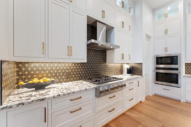 kitchen with light wood-style flooring, light stone counters, appliances with stainless steel finishes, white cabinets, and wall chimney range hood