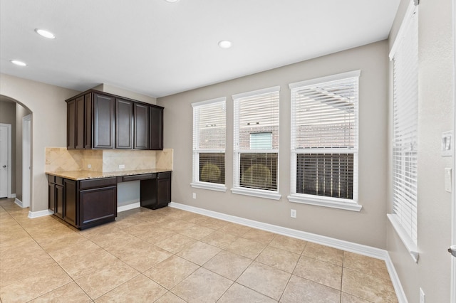 kitchen featuring light stone counters, arched walkways, decorative backsplash, dark brown cabinets, and baseboards