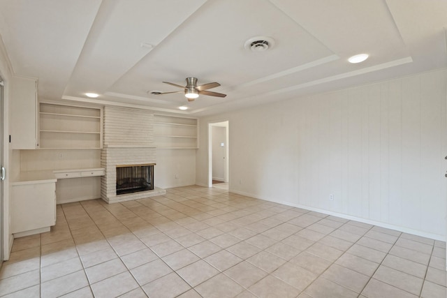 unfurnished living room featuring built in shelves, visible vents, a ceiling fan, a brick fireplace, and a tray ceiling