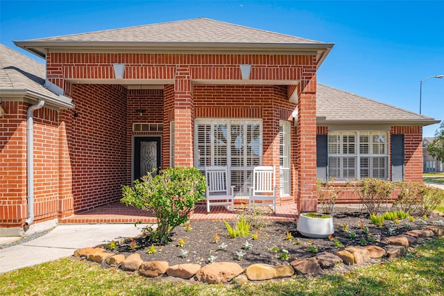 entrance to property featuring brick siding, covered porch, and roof with shingles