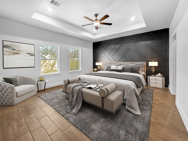 bedroom with a raised ceiling, visible vents, an accent wall, wood finished floors, and baseboards