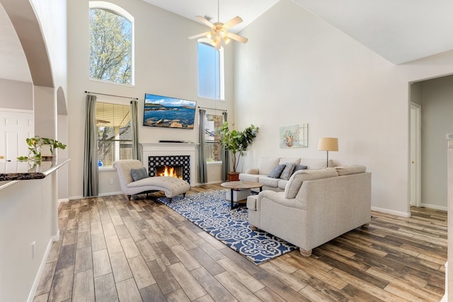 living room with baseboards, a warm lit fireplace, wood finished floors, and a ceiling fan