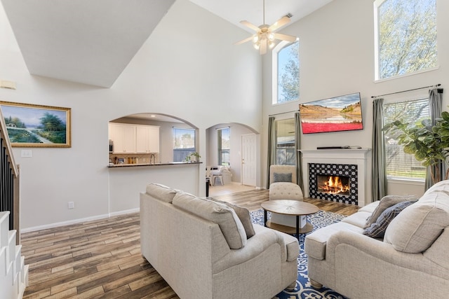 living room featuring ceiling fan, baseboards, a lit fireplace, light wood-style floors, and arched walkways