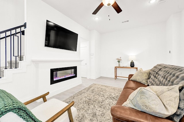 living area featuring visible vents, a glass covered fireplace, light wood-style flooring, ceiling fan, and stairway