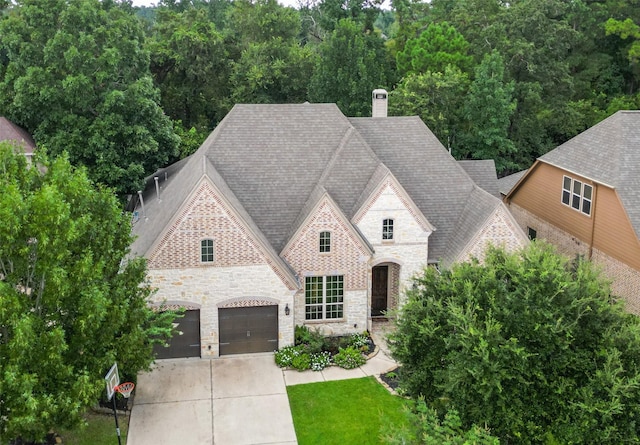 french country home featuring stone siding, roof with shingles, concrete driveway, brick siding, and a chimney