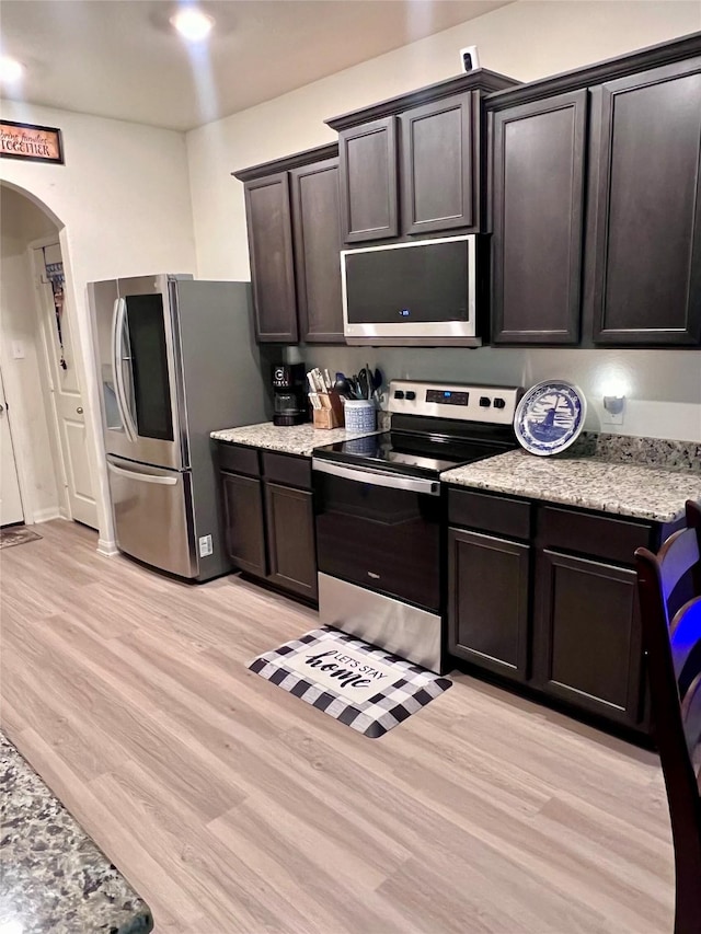 kitchen with light stone counters, stainless steel appliances, arched walkways, dark brown cabinetry, and light wood finished floors