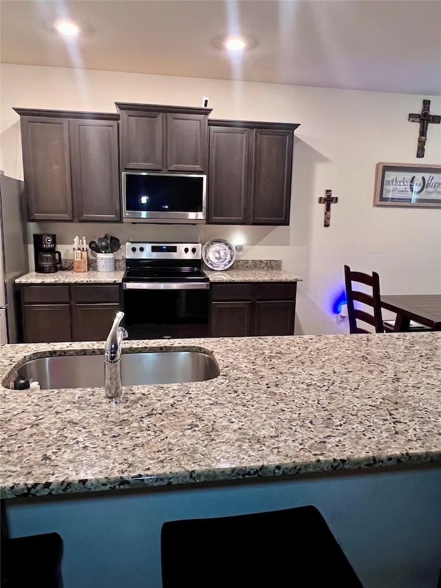 kitchen featuring dark brown cabinets, light stone countertops, stainless steel appliances, and a sink