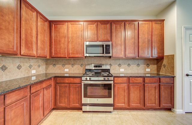 kitchen with backsplash, dark stone counters, light tile patterned floors, brown cabinets, and appliances with stainless steel finishes