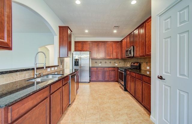 kitchen with dark stone counters, arched walkways, a sink, stainless steel appliances, and tasteful backsplash