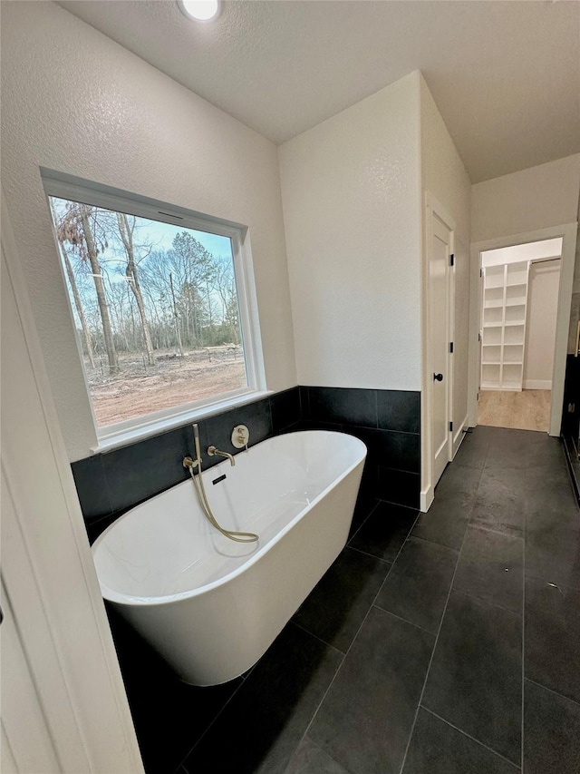 full bathroom with a wainscoted wall, tile patterned floors, a walk in closet, a freestanding tub, and tile walls