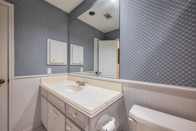 half bath featuring visible vents, wainscoting, toilet, and wallpapered walls