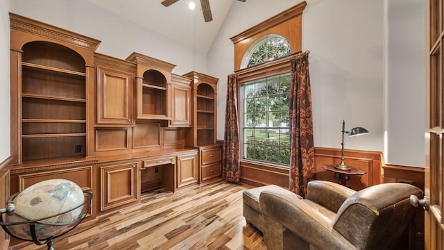 living area with high vaulted ceiling, a ceiling fan, light wood-type flooring, wainscoting, and built in desk